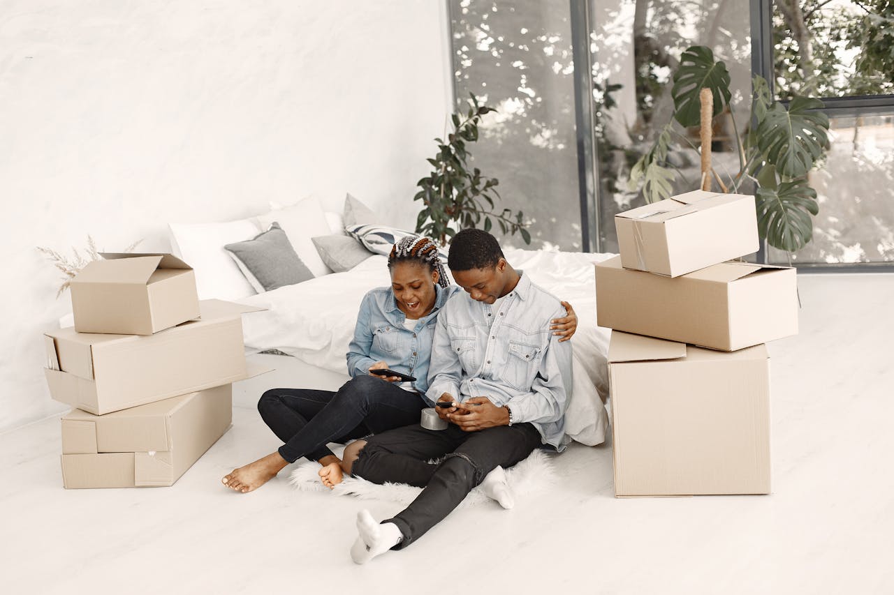 Happy couple sitting with boxes, using smartphones, enjoying moving day indoors.
